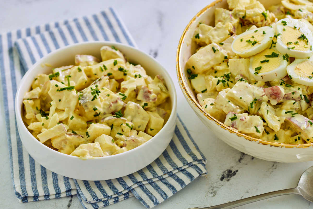 Small bowl of deviled egg potato salad garnished with chives next to large serving bowl with more potato salad topped with hard boiled egg slices.