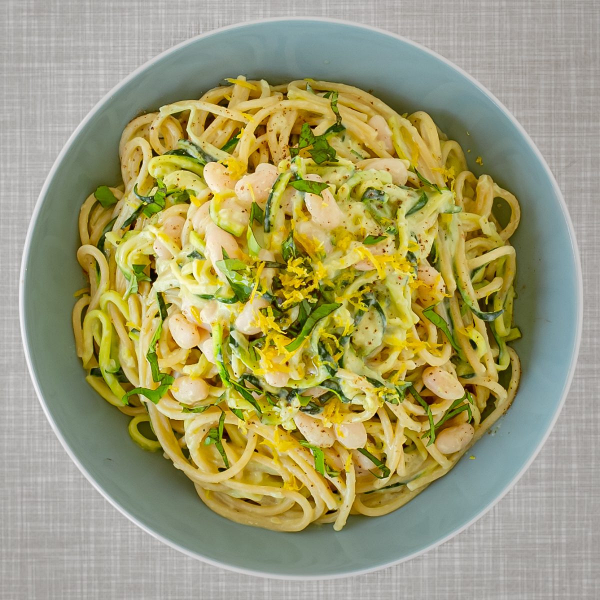 A turquoise plate filled with a vegan creamy lemon zucchini pasta with white beans, and basil. Seen from above