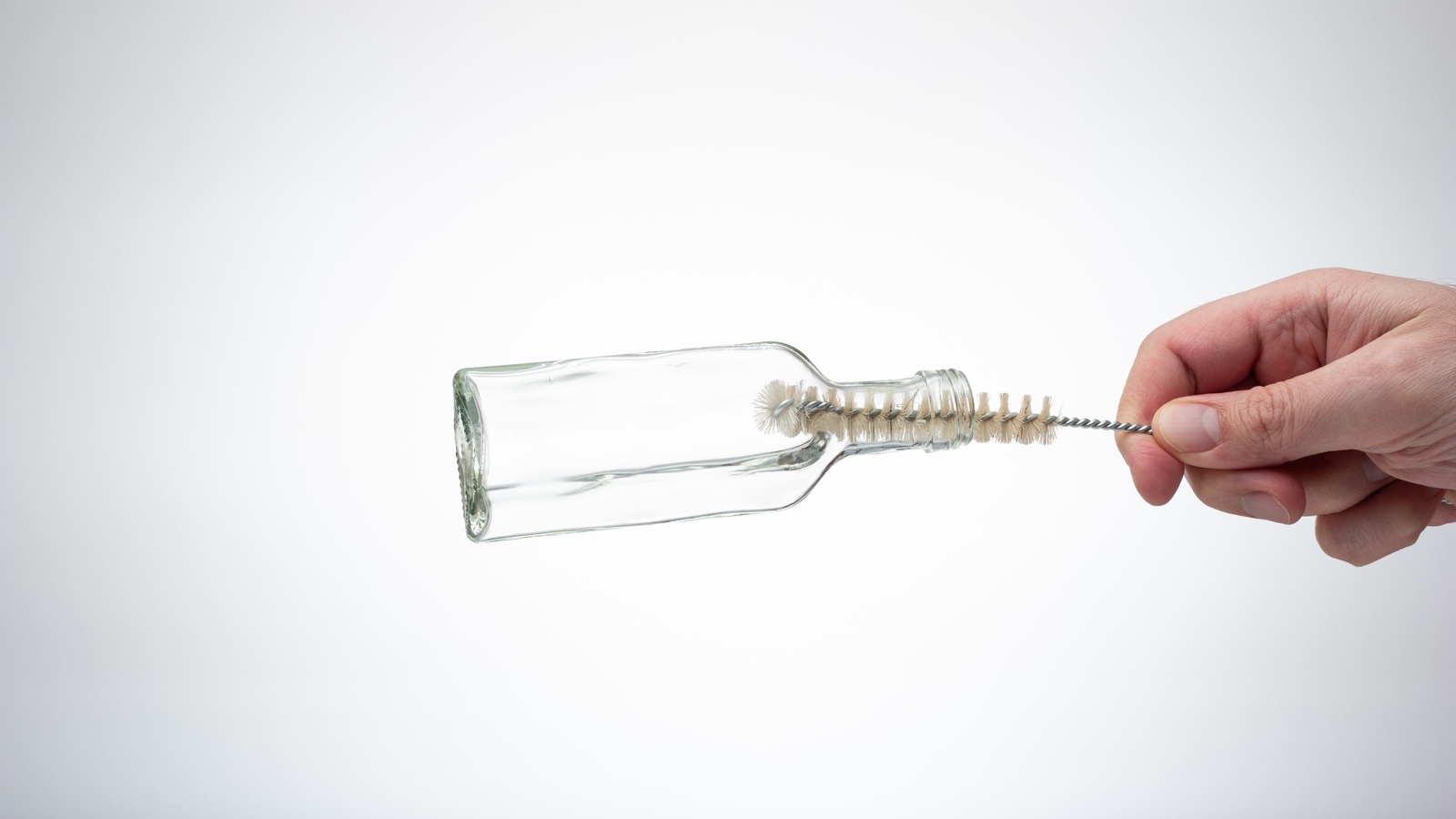 A hand using a small bottle brush to scrub the interior of a clear glass bottle