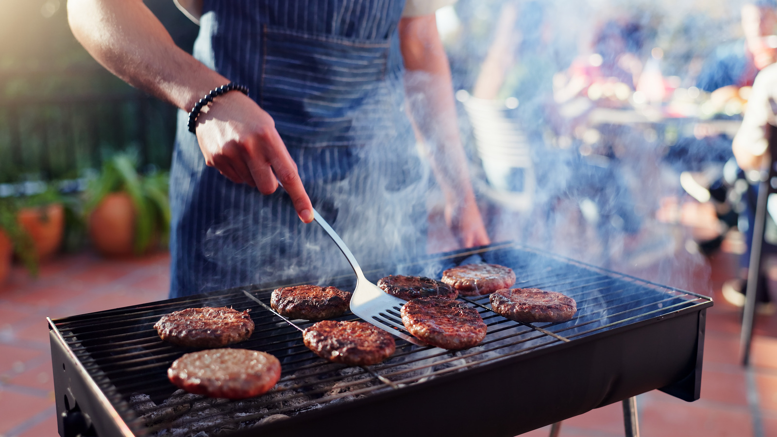 Man using a spatula to cook burgers on a barbecue outdoors on a paved terrace