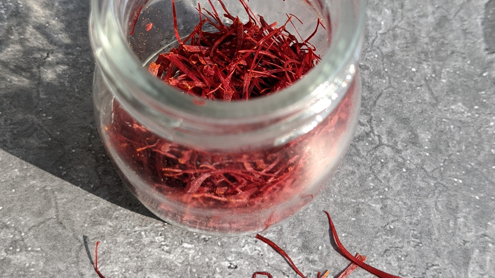 Dried red saffron threads in a small clear glass jar with a few loose strands beside it