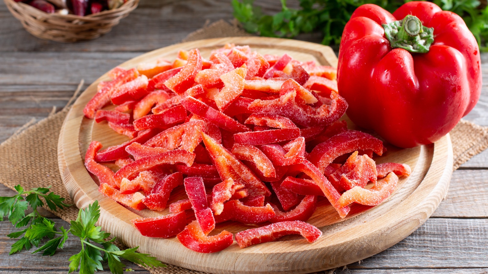 Loose frozen red bell pepper strips on a wooden board beside a fresh pepper