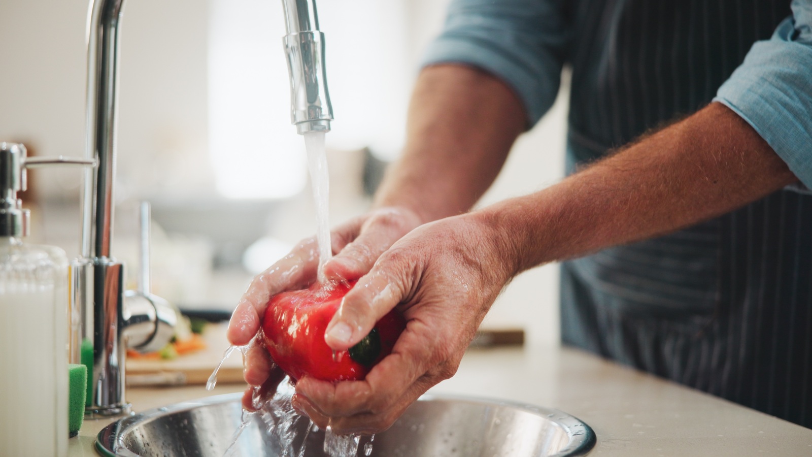 Hands rinsing a whole red bell pepper under running water at a kitchen sink