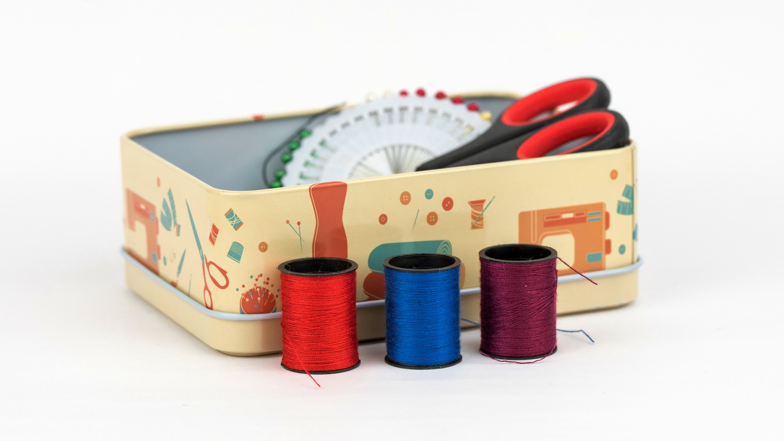 A decorative metal tin holding scissors and sewing supplies with thread spools in front