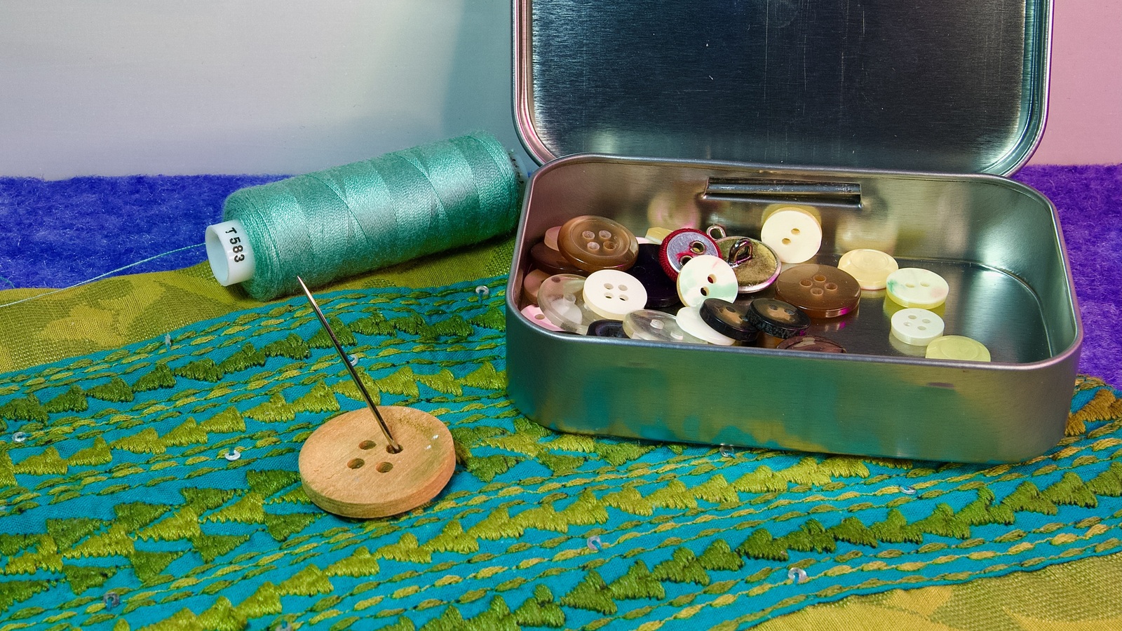 Assorted spare buttons in a metal tin beside a needle and thread