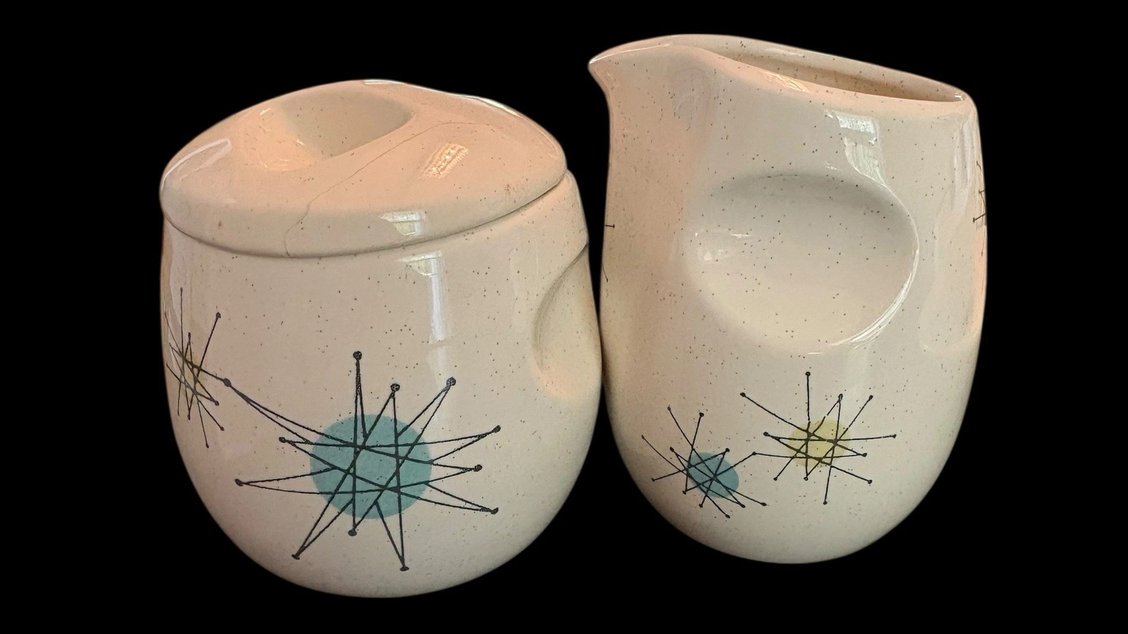 Franciscan Starburst sugar bowl with lid beside a sculpted creamer on a black background.