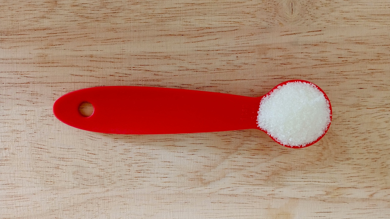 White granulated sugar measured in a red spoon on a wooden surface