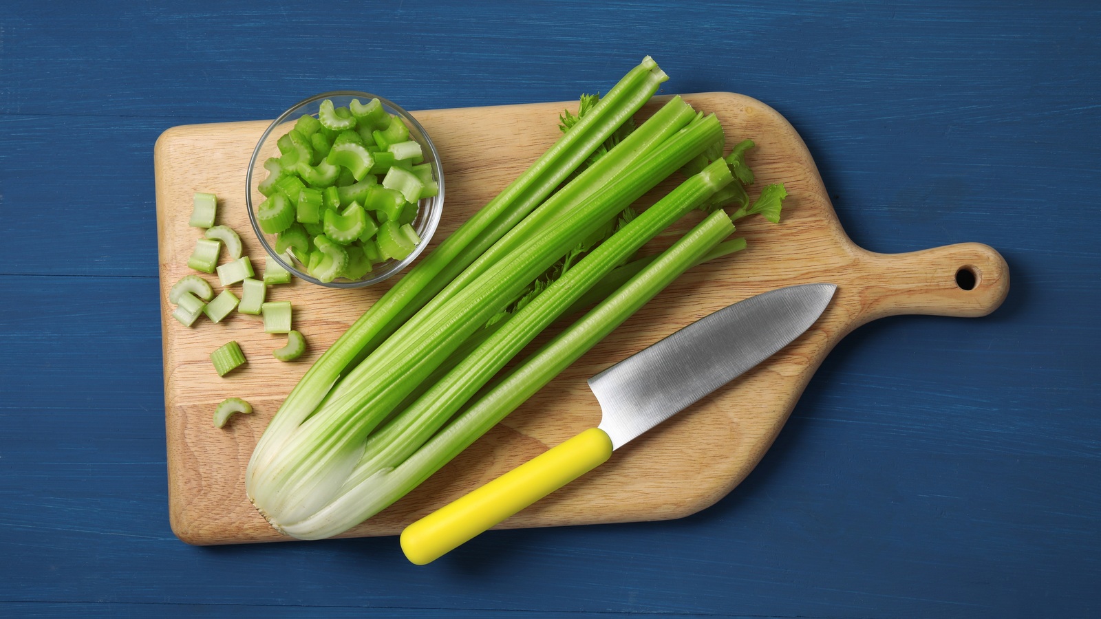 Whole celery stalks, chopped celery in a bowl, and a knife on a wooden board